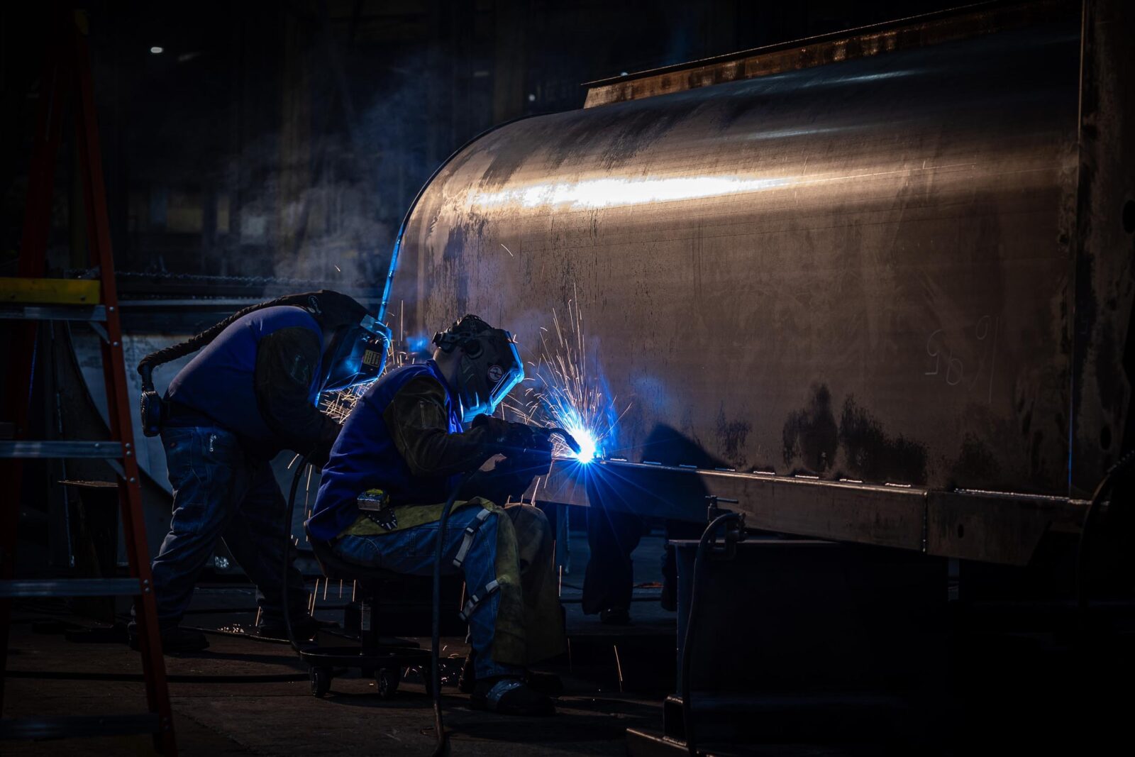 Two people welding on an MTM trailer tub as it's in an upside-down position