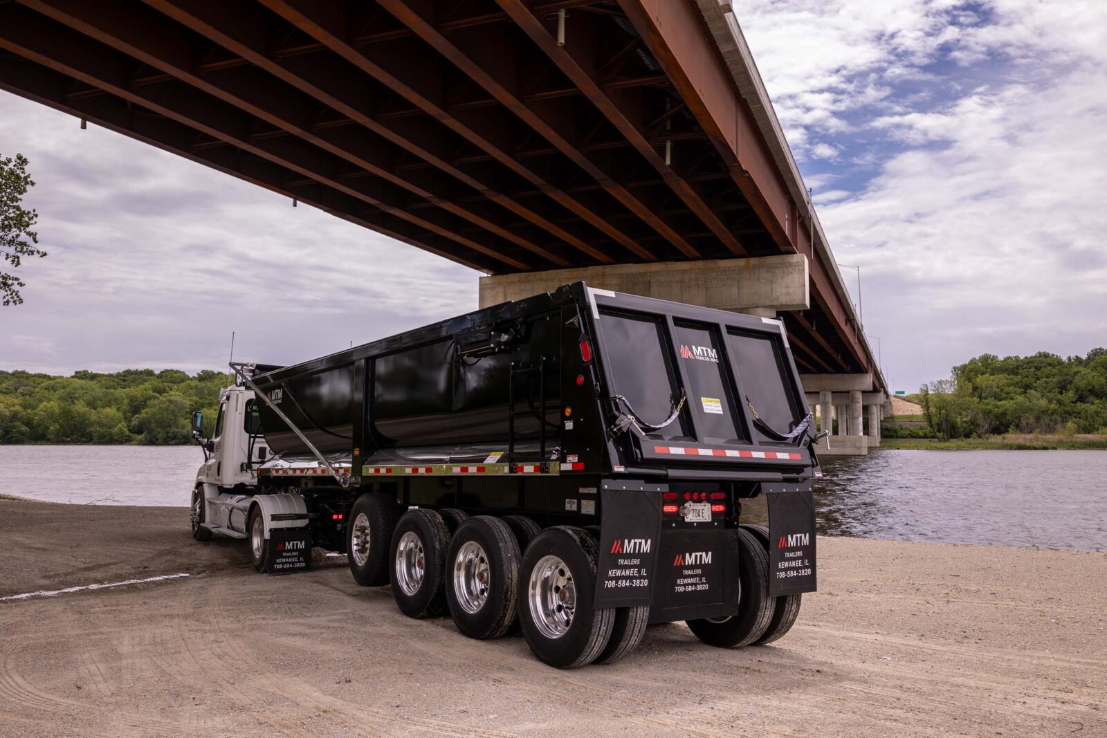 White truck pulling a black quad-axle MTM end-dump trailer with a river in the background, back view