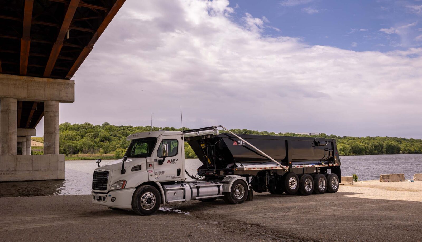 White truck pulling a black quad-axle MTM end-dump trailer with a river in the background