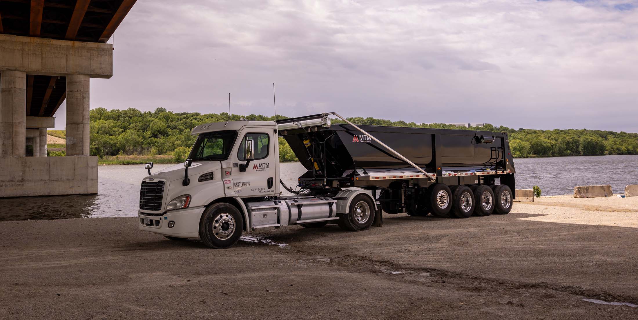 White truck pulling a black quad-axle MTM end-dump trailer with a river in the background